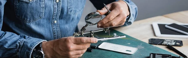 Cropped view of repairman with screwdriver holding magnifier near disassembled part of mobile phone at workplace, banner — Stock Photo