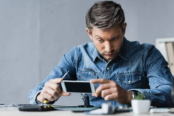 Focused repairman with screwdriver looking through magnifier at mobile phone while sitting at workplace on blurred foreground — Stock Photo