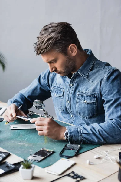 Konzentrierter Reparateur blickt durch die Lupe auf zerlegtes Handy am Arbeitsplatz im verschwommenen Vordergrund — Stockfoto