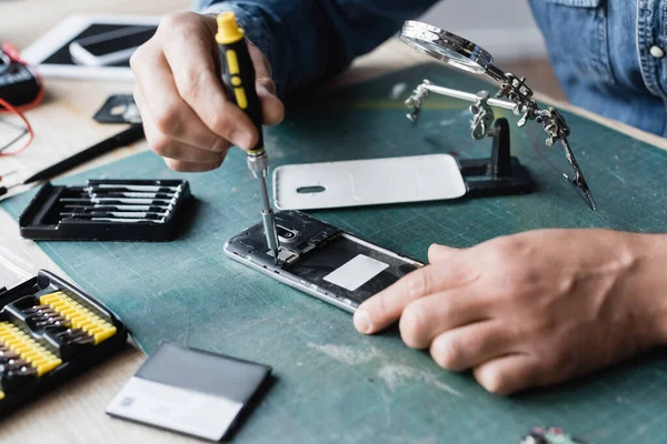 Vista ritagliata del riparatore con cacciavite che fissa il telefono cellulare smontato sul posto di lavoro su sfondo sfocato — Foto stock