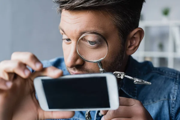 Close up of repairman looking through magnifier at cellphone with blank screen on blurred foreground — Stock Photo
