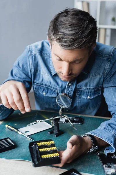 Reparateur blickt auf Halterung mit Schraubendreher-Bits am Arbeitsplatz mit zerlegten Mobiltelefonen auf verschwommenem Hintergrund — Stockfoto