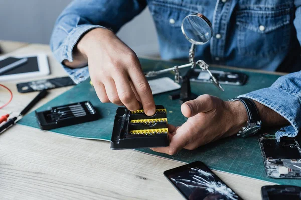 Ausgeschnittene Ansicht eines Reparateurs, der Schraubendreher-Bit aus Halterung nimmt, während er am Arbeitsplatz auf verschwommenem Hintergrund sitzt — Stockfoto