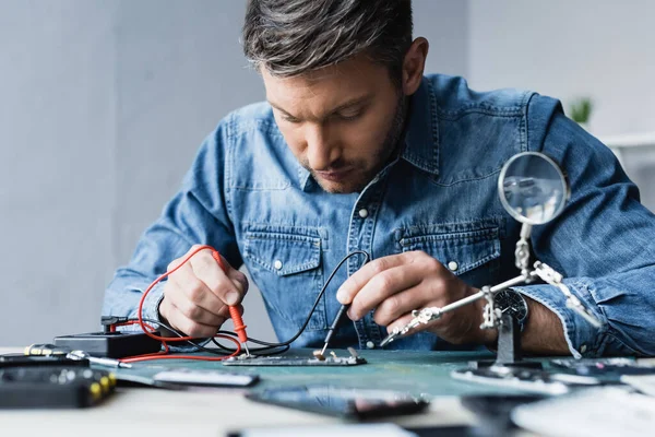 Konzentrierter Reparateur mit Multimeter-Sensoren auf zerlegtem Teil des Mobiltelefons im verschwommenen Vordergrund — Stockfoto