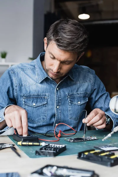 Riparatore focalizzato che regola il multimetro tenendo premuto il sensore su una parte smontata del telefono cellulare in primo piano sfocato — Foto stock