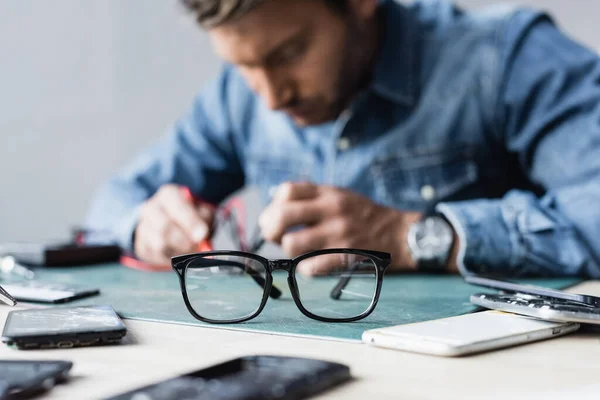 Lunettes de vue sur table près de pièces démontées de téléphones mobiles avec réparateur flou travaillant sur fond — Photo de stock