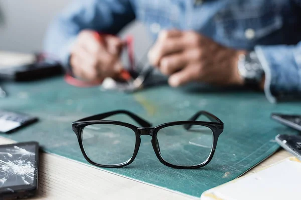 Nahaufnahme der Brille mit schwarzem Rahmen auf dem Tisch in der Nähe von kaputten Mobiltelefonen mit verschwommenem Werkstattmitarbeiter auf dem Hintergrund — Stockfoto