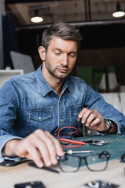 Serious repairman holding hand near eyeglasses on table with disassembled parts of mobile phone on blurred foreground — Stock Photo