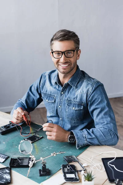 Happy repairman looking at camera with sensors of multimeter while sitting at table with disassembled parts of smartphones — Stock Photo