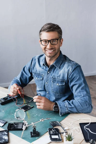 Réparateur heureux regardant la caméra avec des capteurs de multimètre tout en étant assis à table avec des pièces démontées de smartphones — Photo de stock