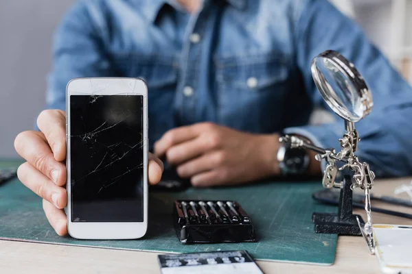 Vista recortada del reparador que muestra el teléfono celular roto mientras está sentado en la mesa cerca de la lupa y el kit de herramientas en un fondo borroso - foto de stock