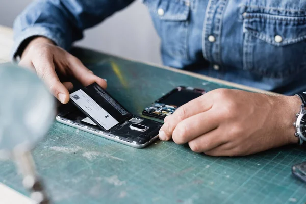 Ausgeschnittene Ansicht eines Reparateurs, der Akku in Handy auf Tisch legt, mit verschwommener Lupe im Vordergrund — Stockfoto