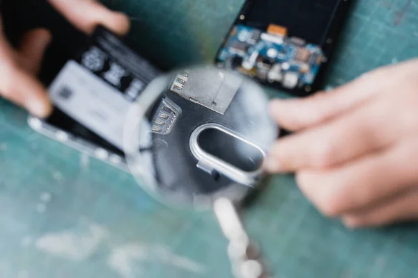 Cropped view of repairman holding magnifier over broken parts of mobile phones on blurred foreground — Stock Photo