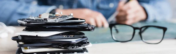 Close up view of pile of broken mobile phones near eyeglasses on workplace with blurred repairman on background, banner — Stock Photo