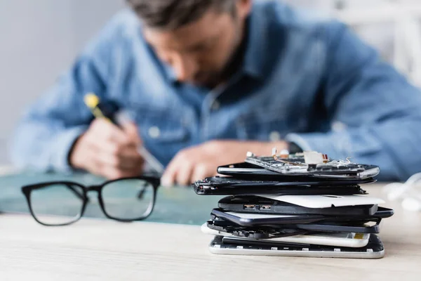 Close up view of pile of broken mobile phones near eyeglasses on workplace with blurred repairman on background — Stock Photo