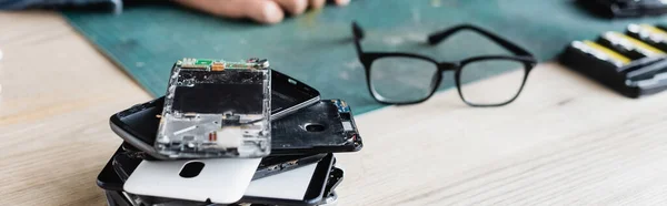 Nahaufnahme eines Haufens zerbrochener Mobiltelefone in der Nähe von Brillen am Arbeitsplatz mit verschwommenem Werkstattmitarbeiter im Hintergrund, Banner — Stockfoto