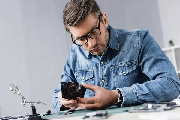 Reparador enfocado desmontar el teléfono inteligente roto mientras está sentado en el lugar de trabajo con lupa en primer plano borroso - foto de stock