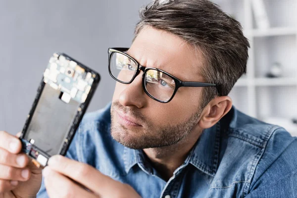 Réparateur concentré dans des lunettes regardant une partie démontée du téléphone mobile au premier plan flou — Photo de stock