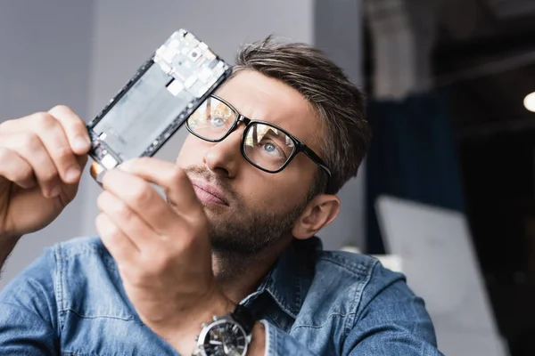 Reparador focado em óculos olhando para o componente desmontado do telefone móvel em fundo embaçado — Fotografia de Stock