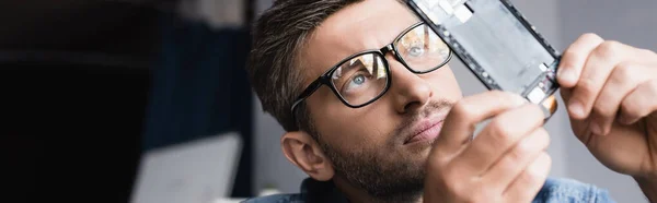 Focused repairman in eyeglasses looking at disassembled part of broken mobile phone on blurred background, banner — Stock Photo