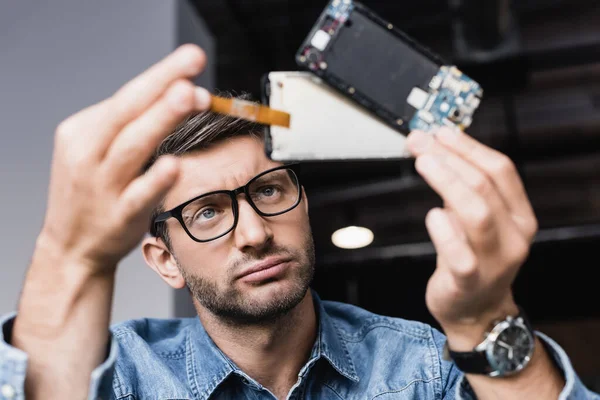 Dissatisfied repairman in eyeglasses looking at disassembled parts of broken mobile phone on blurred foreground — Stock Photo