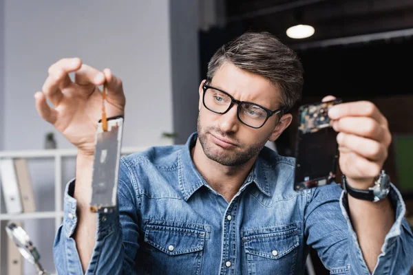 Skeptical repairman in eyeglasses holding disassembled parts of broken smartphone with blurred magnifier on foreground — Stock Photo