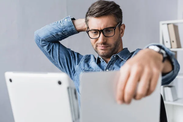 Reparador cuidadoso com a mão atrás da cabeça olhando para laptop danificado em primeiro plano desfocado — Stock Photo