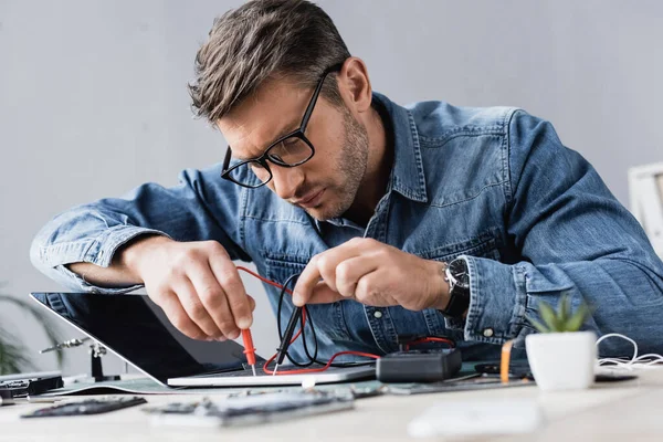 Focused repairman holding sensors of multimeter on broken keyboard of laptop at workplace on blurred foreground — Stock Photo