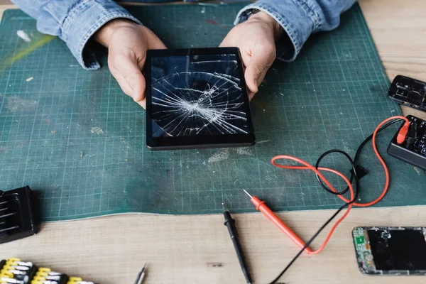 Vue recadrée du réparateur tenant une tablette numérique fracassée sur le lieu de travail près du multimètre et des téléphones mobiles démontés — Photo de stock