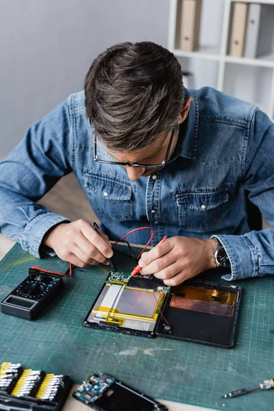 Repairman with holding sensors of multimeter on broken part of digital tablet at workplace — Stock Photo