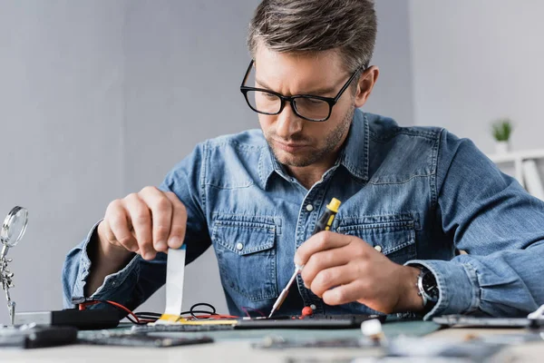 Réparateur focalisé avec tournevis tenant le câble flexible de la tablette numérique démontée avec lieu de travail flou au premier plan — Photo de stock