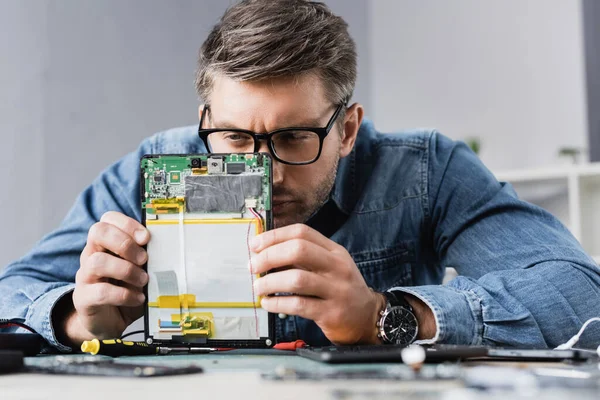 Сосредоточенный ремонтник щурясь, смотрит на часть сломанного цифрового планшета с размытым рабочим местом на переднем плане — стоковое фото