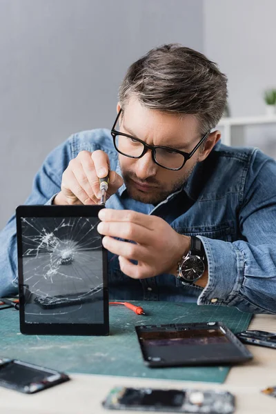 Focused repairman with screwdriver fixing display of smashed tablet with blurred workplace on foreground — Stock Photo