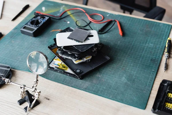 Hochwinkelaufnahme eines Haufens zerlegter Mobiltelefone am Arbeitsplatz mit Schraubenzieher, Lupe und Multimeter — Stockfoto