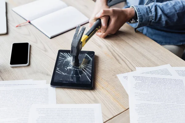 Vue recadrée de l'homme d'affaires frappant tablette numérique avec le marteau tout en étant assis sur le lieu de travail avec des documents — Photo de stock