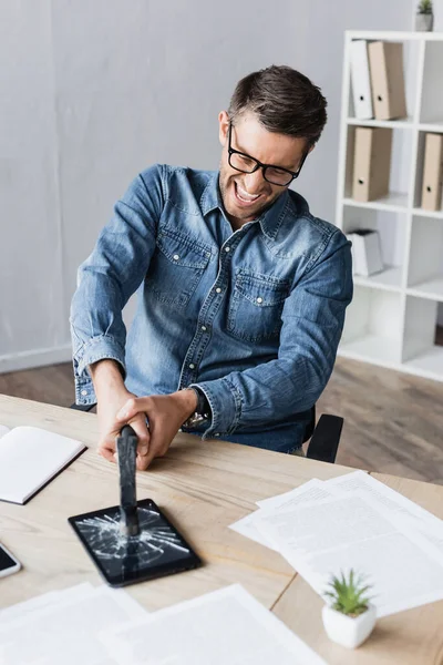 Uomo d'affari arrabbiato in occhiali che colpisce tablet digitale con martello mentre seduto sul posto di lavoro in primo piano sfocato — Foto stock