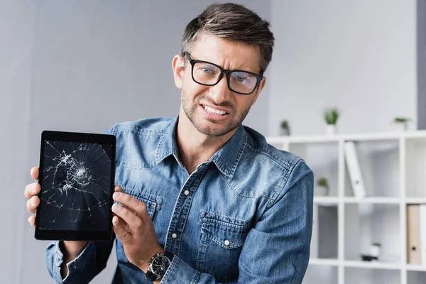 Homme d'affaires en colère dans les lunettes avec tablette numérique fracassée regardant la caméra sur fond flou — Photo de stock