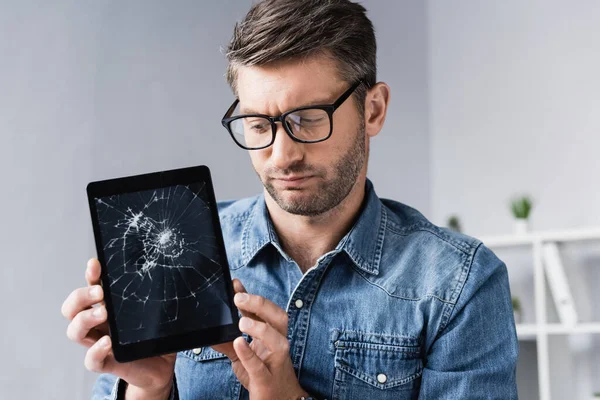 Dissatisfied businessman in eyeglasses looking at smashed digital tablet on blurred background — Stock Photo