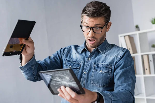 Uomo d'affari sorpreso con la bocca aperta guardando smontato fracassato touchscreen del tablet digitale su sfondo sfocato — Foto stock