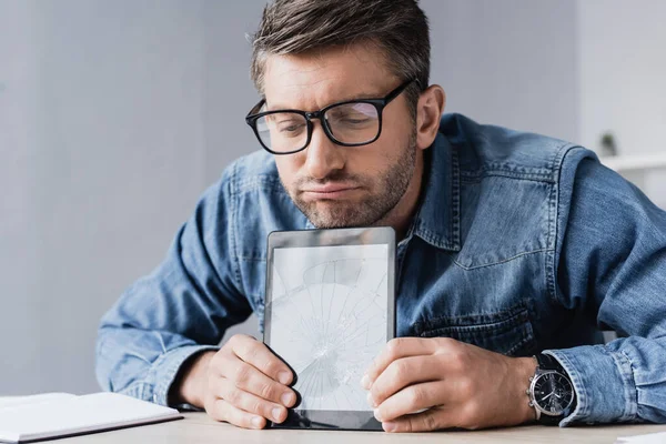 Upset businessman in eyeglasses holding smashed digital tablet while sitting at table — Stock Photo