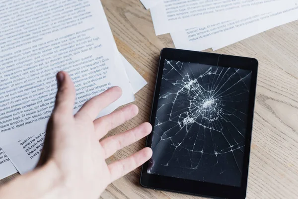 Cropped view of man with hand near smashed digital tablet and documents on wooden background — Stock Photo