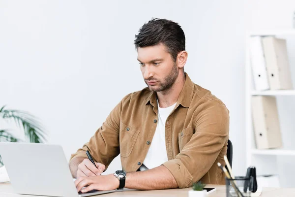 Сосредоточенный бизнесмен с ручкой смотрит на ноутбук, сидя на рабочем месте на размытом фоне — стоковое фото