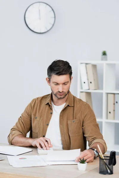 Hombre de negocios enfocado mirando la hoja de papel mientras cuenta con calculadora en el lugar de trabajo sobre fondo borroso - foto de stock