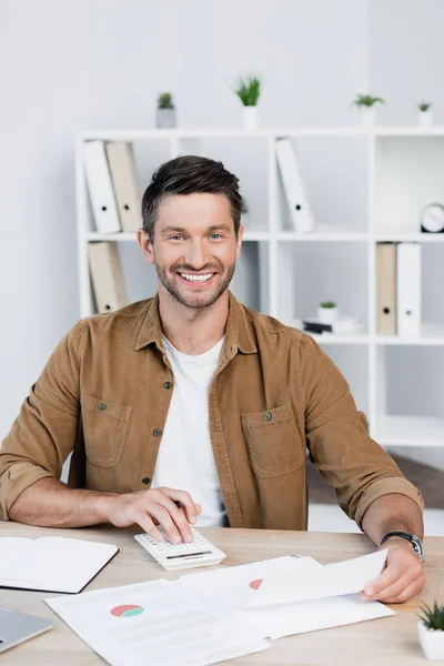 Cheerful businessman looking at camera while counting on calculator at workplace on blurred background — Stock Photo