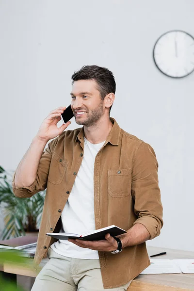 Cheerful businessman with notebook talking on mobile phone while leaning on table on blurred background — Stock Photo