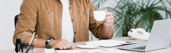 Vista cortada de empresário com xícara de café sentado no local de trabalho com laptop com planta turva no fundo, banner — Fotografia de Stock