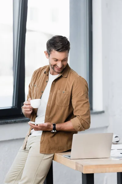Empresário sorridente com xícara de café e pires olhando para laptop enquanto se inclina na mesa com janela borrada no fundo — Fotografia de Stock