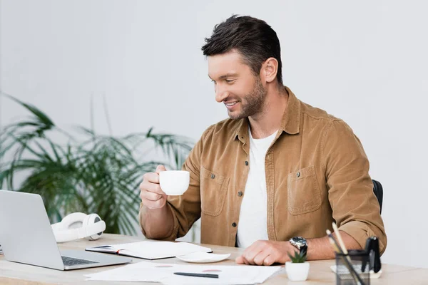 Cheerful businessman with coffee cup looking at laptop while sitting at workplace with blurred plant on background — Stock Photo