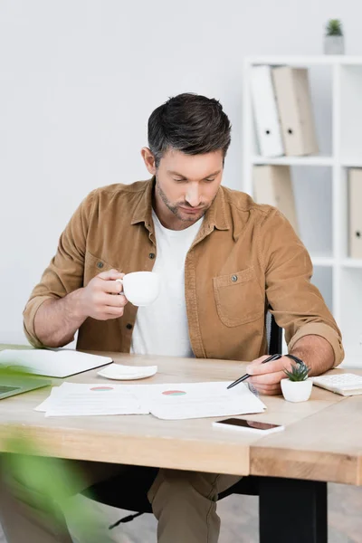 Focused businessman with coffee cup pointing with pen at paper sheet while sitting at workplace on blurred foreground — Stock Photo