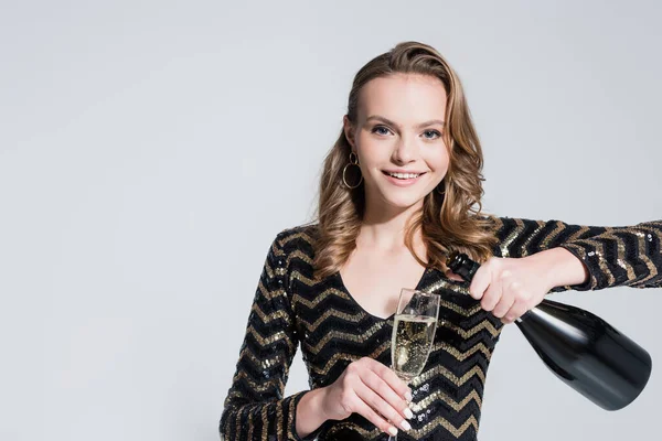 Femme heureuse tenant verre tout en versant du champagne de bouteille isolé sur gris — Photo de stock